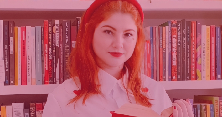 Foto da Tayane Gomes, a Médica Leitora, ao fundo. Uma mulher branca, com cabelos ruivos